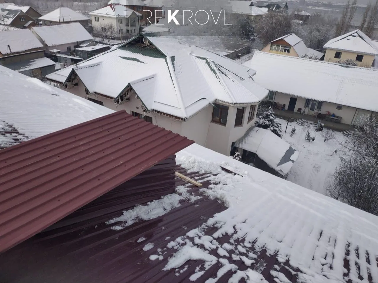 Вид с крыши: бордовый профнастил под снегом и заснеженные соседние дома