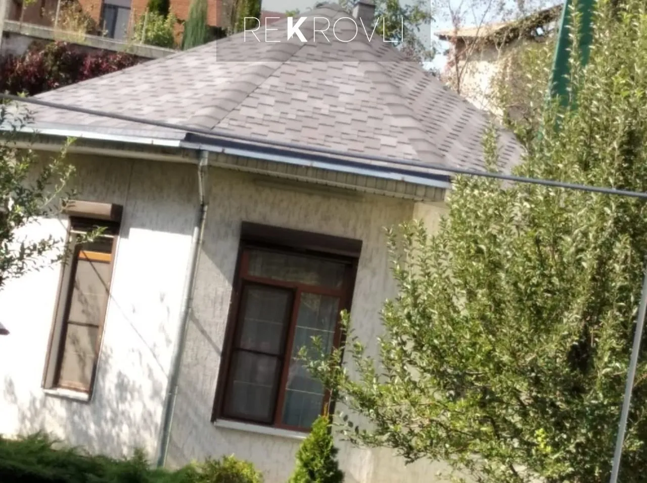 Шатровая крыша из серой гибкой черепицы на одноэтажном частном доме среди деревьев